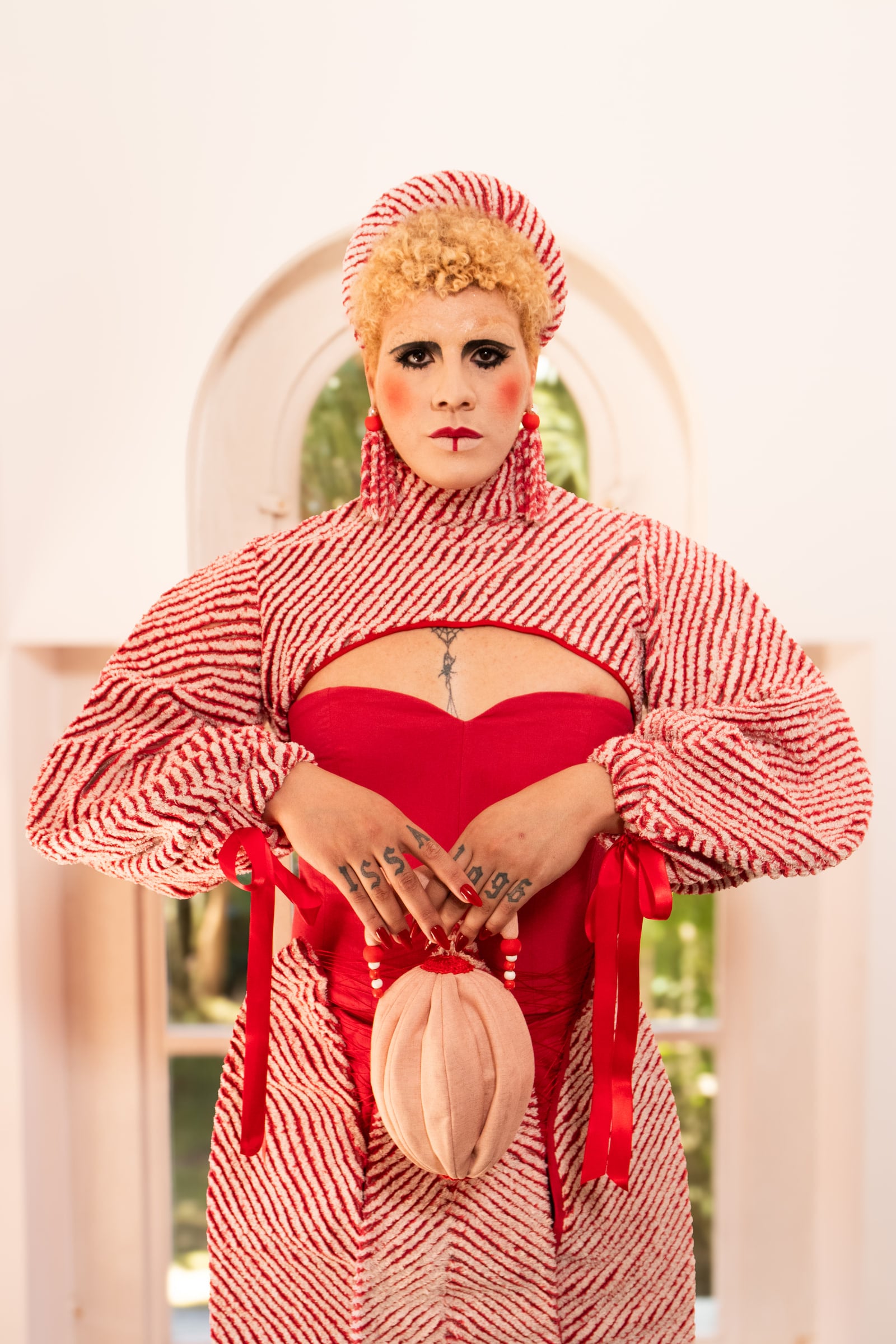 Modelo de frente diante de uma janela em arco, com as mãos sobre um acessório oval claro e o vestido vermelho sob a camada listrada.