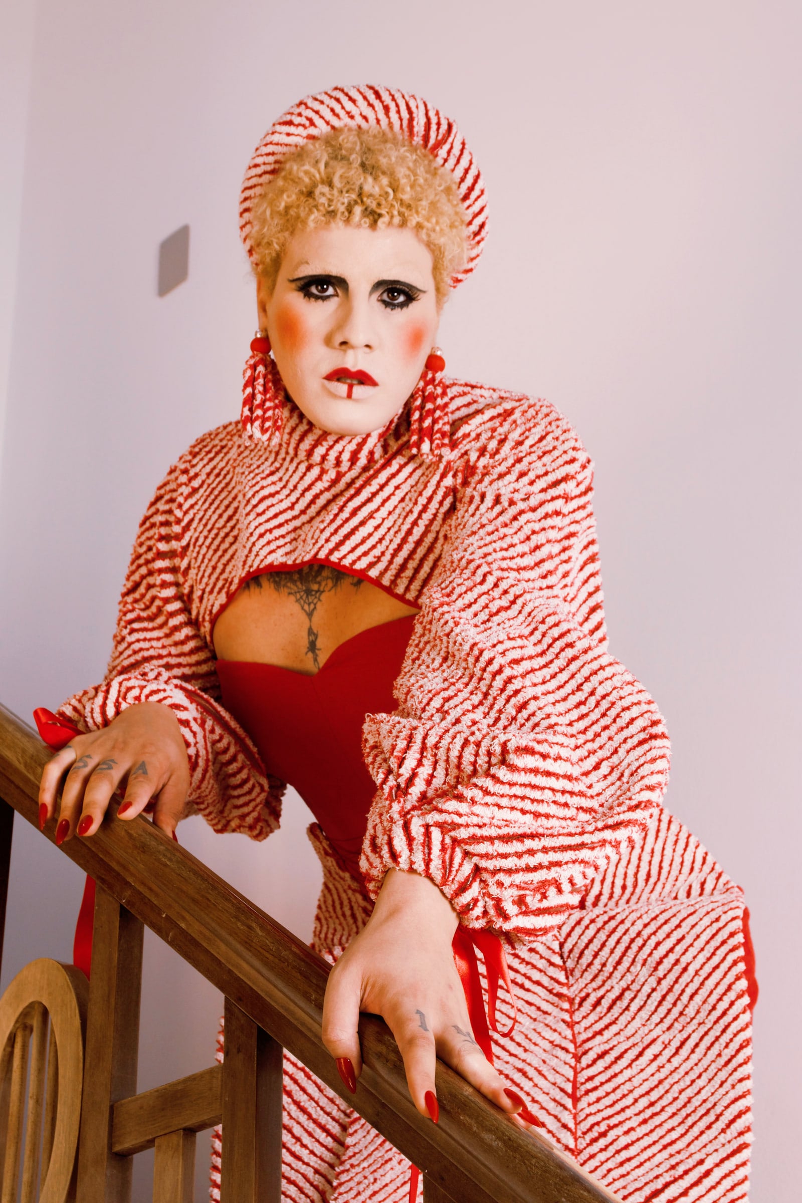 Modelo apoia as mãos no corrimão de madeira. A peça listrada envolve o pescoço e os ombros sobre um vestido vermelho.